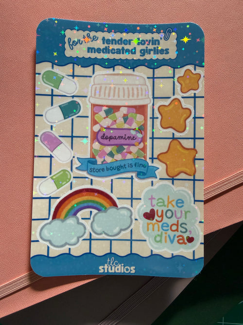 A rectangular sticker sheet with rounded corners featuring colorful cartoon medicine-themed designs, including pills, a pill bottle labeled 'dopamine,' stars, a rainbow, clouds, and phrases like 'take your meds diva.' The sheet is photographed on a stack of journals.